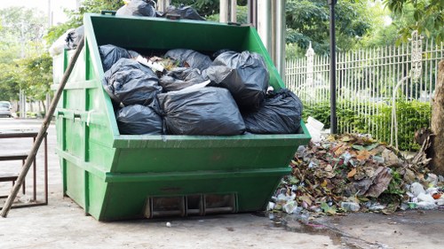A large green skip bin filled with black rubbish bags sits on a paved outdoor area near a park or garden, with some fallen autumn leaves and scattered waste visible on the ground beside it. The skip is positioned next to a metal fence and a tree, with natural light illuminating the scene. The environment suggests an urban or suburban setting, likely in Woking, where House Clearance Woking offers rubbish removal services. The skip's textured surface appears weathered, and the black bags are tightly packed, indicating the collection of household or garden waste. The scene captures a typical rubbish disposal point, with the surrounding greenery and nearby fencing enhancing the clear boundaries of the waste management area, reflecting the practical aspects of domestic or commercial clearance projects in the local area.