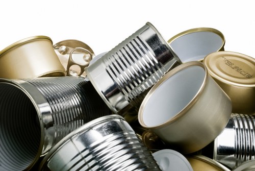 A close-up image displaying a collection of used metal cans, primarily made of tinplate with a shiny, reflective surface. The cans are cylindrical and vary in size, with some featuring pull-tabs on their lids while others have smooth, closed tops. They are arranged in a haphazard pile, with some leaning against each other and others lying flat, on an unseen surface. The background is neutral and blurred, emphasizing the metallic textures and tones of silver and gold. This arrangement is typical of waste collection or rubbish associated with household or commercial waste disposal, which House Clearance Woking might manage during rubbish removal services in the Woking area, suggesting an environment involving the handling of metal recyclable waste materials.