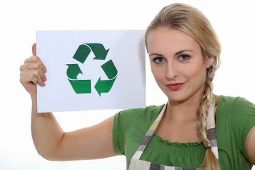 A young woman with blonde hair styled in a side braid, wearing a green t-shirt and a striped apron, is holding a white sign with a green recycling symbol consisting of three arrows forming a triangle, in front of a plain white background. The woman is facing forward and has a neutral facial expression. The image emphasizes environmental themes and responsible waste management, aligning with services offered by companies like House Clearance Woking. The recycling symbol on the sign highlights environmentally conscious rubbish removal or house clearance practices, and the simple, clean background keeps the focus on the woman and the sign. The visual presentation suggests a professional and approachable attitude towards waste disposal and recycling services in the Woking area, subtly supporting local SEO without explicit promotional language.