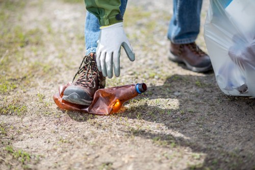A person wearing blue jeans, green jacket, and white gloves is picking up a used glass bottle from a dirt ground, possibly during a cleanup activity. The bottle, which is brown with an open mouth, is positioned near the person's ankle. In the background, part of a brown boot and a grey plastic bag are visible, indicating an outdoor setting such as a garden or yard. The ground is a mixture of soil and small grass patches, with natural lighting suggesting daytime. The scene appears to be part of a rubbish removal or litter collection effort, reflecting environmental care and waste management practices. This type of activity aligns with services offered by House Clearance Woking, specializing in rubbish removal and clearance tasks in the local area. The environment looks tidy apart from the discarded bottle, emphasizing the importance of proper waste disposal and community cleanliness.