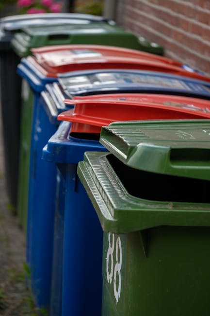 A close-up view of crushed and wrinkled aluminium beverage cans, predominantly silver in colour with some featuring faded branding such as Coca-Cola in red and white. The cans exhibit smooth, reflecti