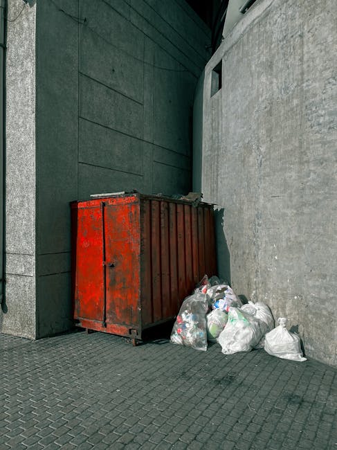 A rusty red metal waste container, situated in a narrow alleyway corner between a dark green wall on the left and a light grey concrete wall on the right, is partially covered in peeling paint and sho
